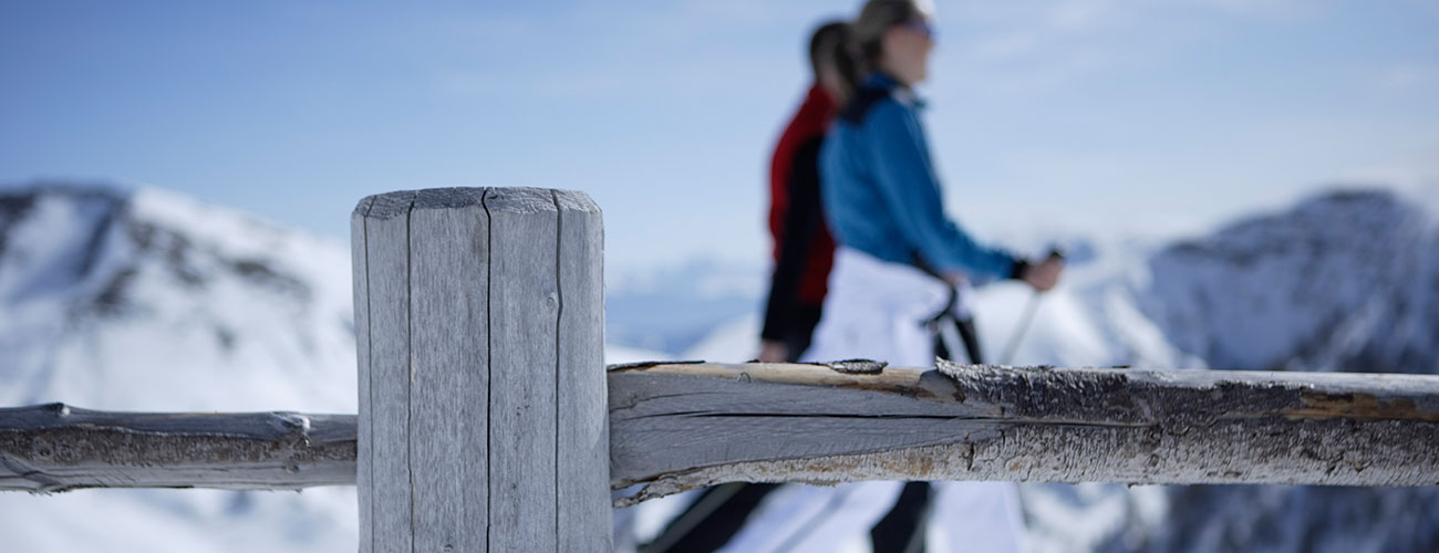 Detailaufnahme eines Holzzauns mit zwei Winterwanderern und Schneelandschaft im Hintergrund