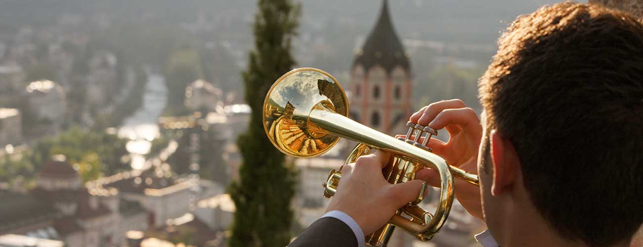 Mann bläst Trompete mit der Stadt Meran im Hintergrund